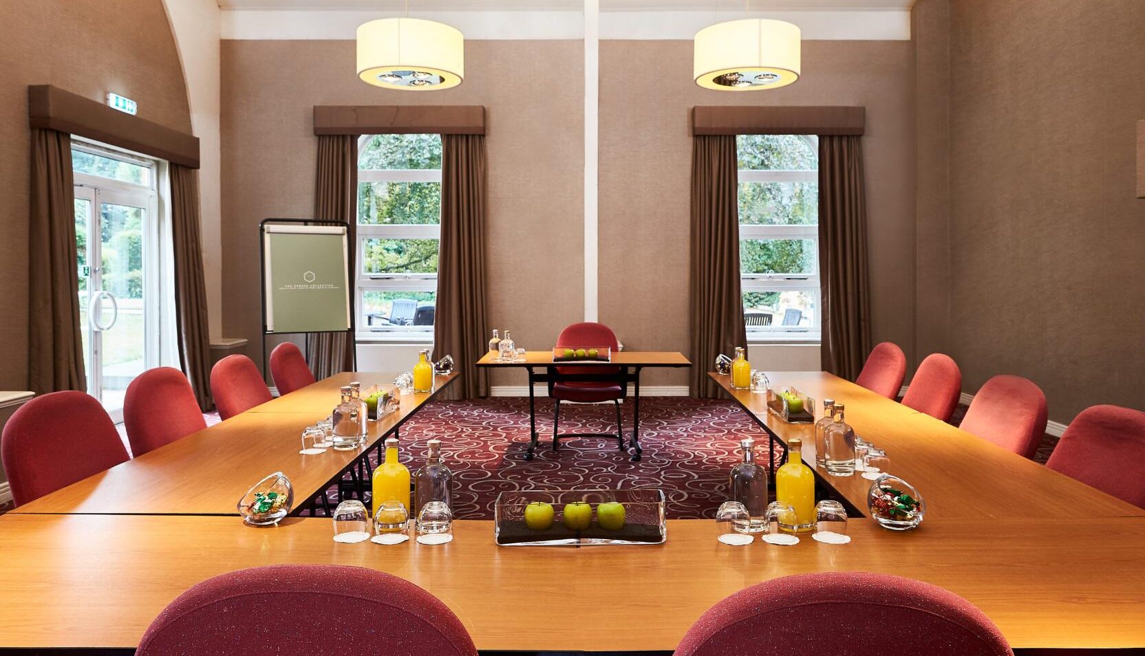 Conference room with red chairs, wooden tables set with glasses, glass water, a juice bottle, green apples, and sweets in a glass bowl.